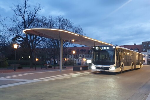 Ein Bus steht an einem Steig des Bernburger Busbahnhofs. Das eliptische Dach des der Station ist beleuchtet. Auch der Bus steht mit angeschalteten Lichtern. 