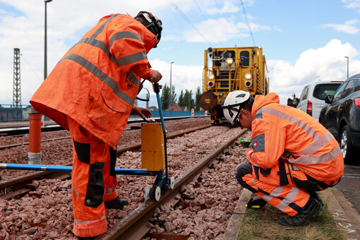 Arbeiten am Gleis einer Straßenahn. Zwei Bauarbeiter in signal-orange-farbener Berufsbekleidung arbeiten an den Schienen mit einer sogenannten Stopfmaschine. 