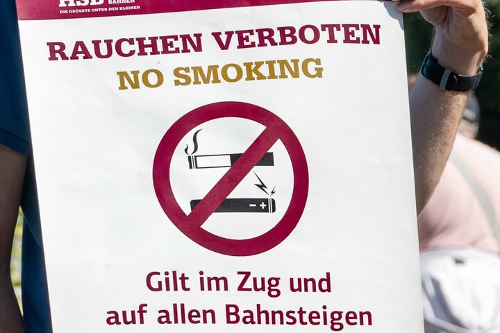 Eine Person, deren Kopf nicht im Bildausschnitt zu sehen ist, hält ein weißes Verbotsschild vor dem Oberkörper. Auf dem Schild sind eine Zigarette und eine E-Zigarette abgebildet, beide durchgestrichen in einem weinroten Kreis mit Querlinie – dem klassischen Symbol für ein Rauchverbot. Darüber steht in großer weinroter Schrift: „Rauchen verboten“. Darunter folgen zwei weitere Hinweise: „Gilt im Zug und auf allen Bahnsteigen“ sowie „Bei Verstoß 250 Euro Bußgeld“. Unter jedem dieser Sätze befindet sich die englische Übersetzung in khakigrüner Schrift: „No Smoking“, „In trains and on all platforms“ sowie „Fine of 250€ for violations“. Die Gestaltung ist schlicht, mit Fokus auf den Warnhinweis.