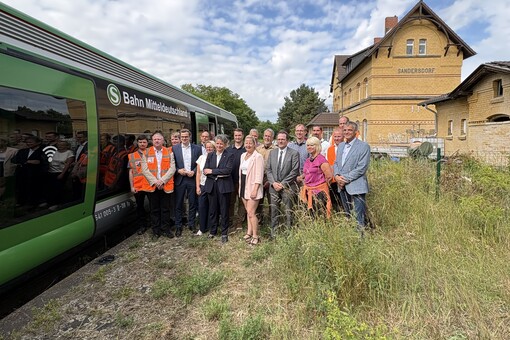Gruppenbild, unter anderem mit Ministerin Hüskens und Staatssekretär Haller, an einem zugewachsenen Bahnsteig. Links neben der Gruppe steht ein grün-weißer Zug. Rechts ist ein Bahnhofsgebäude mit der Aufschrift Sandersdorf zu sehen.