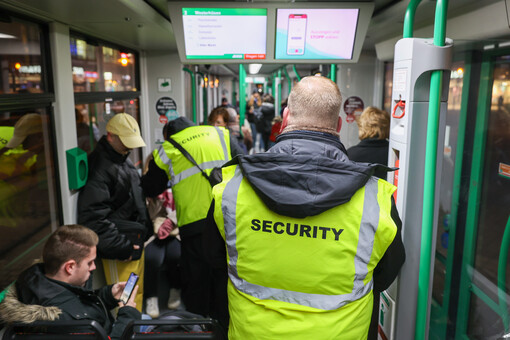 In einer vollbesetzten Straßenbahn stehen zwei Personen mit dem Rücken zu uns. Sie tragen Reflex-Westen mit der Aufschrift Security.