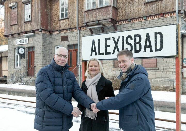 Zwei Männer und eine Frau (Markus Hercher, HSB-Geschäftsführerin Katrin Müller und Harzgerodes Bürgermeister Marcus Weise) geben sic vor einem Bahnhoffschild mit der Auschrift "Alexisbad" die Hand