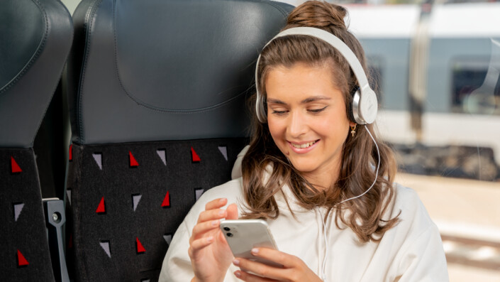 Eine Junge Frau sitzt im Zug und schaut froh auf ihr Handy.